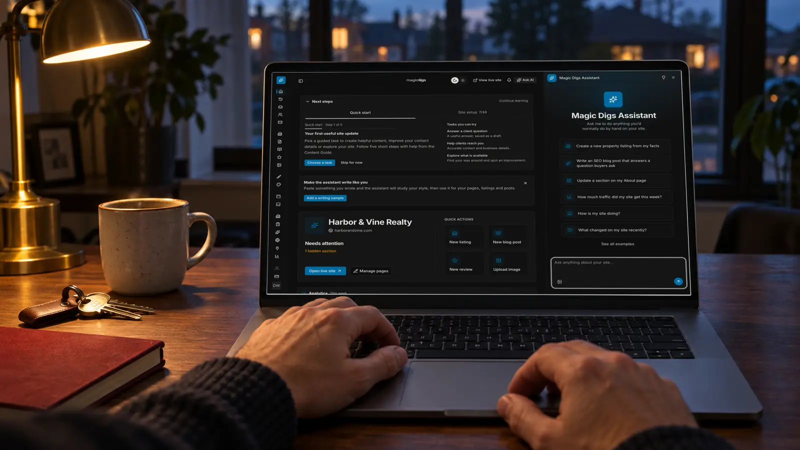 An open laptop on a desk at dusk showing the Magic Digs dashboard with the assistant panel open, with a mug, keys and a notebook beside it